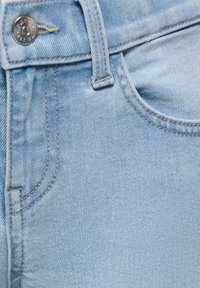 Világoskék farmernadrág közeli kép, amelyen látható az elülső zseb, a derékpánt és az ezüst gomb látható varrási részletekkel.