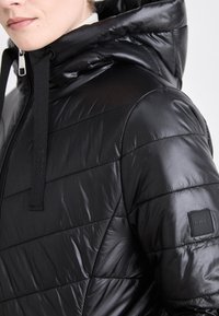 Svart puffjakke med glansfull overflate, horisontal polstring og hette. Har glidelås og et logo-patch på ermet.