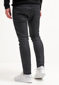 Fekete, slim fit farmernadrág sima textúrával, hátsó zsebekkel és ezüst díszítéssel. Fehér sportcipővel viselve.
