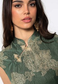 Top de tela verde con un acabado texturizado, que presenta patrones florales dorados y broches decorativos en el escote. Cabello largo y suelto y pendientes.