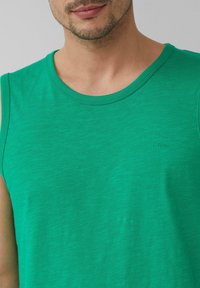 Grünes Tanktop mit Rundhalsausschnitt und einer weichen, glatten Textur. Es zeigt ein dezentes Logo in der unteren linken Ecke.