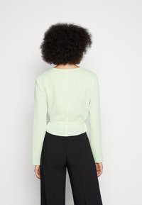Blusa de manga longa verde claro com decote redondo, laço na cintura e parte de trás ajustada. Combinada com calças largas pretas. Textura suave.