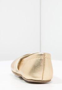 Gull metallisk flatt sko med rund tå, mykt lær overdel og elastisk stropp. Semsket-liknende såle med en sømdetalj ved hælen.
