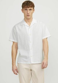 Cămașă din in alb cu mânecă scurtă, cu guler spread, având nasturi deschiși la culoare și o croială lejeră, purtată cu pantaloni crem.