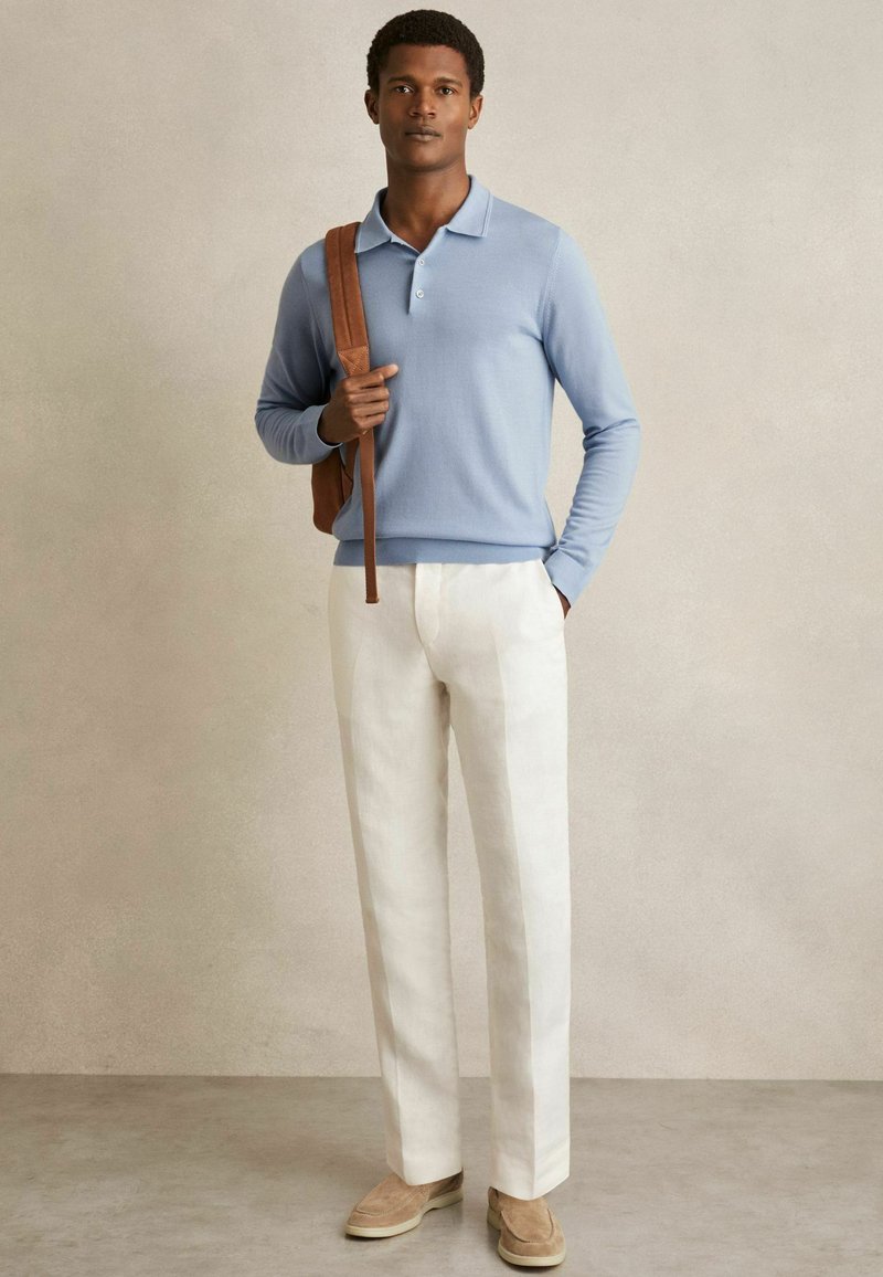 Hellblaue Langarm-Polohemd mit zwei Knöpfen, kombiniert mit beigen Leinenhosen und braunen Wildlederslippern. Braune Lederhandtasche.