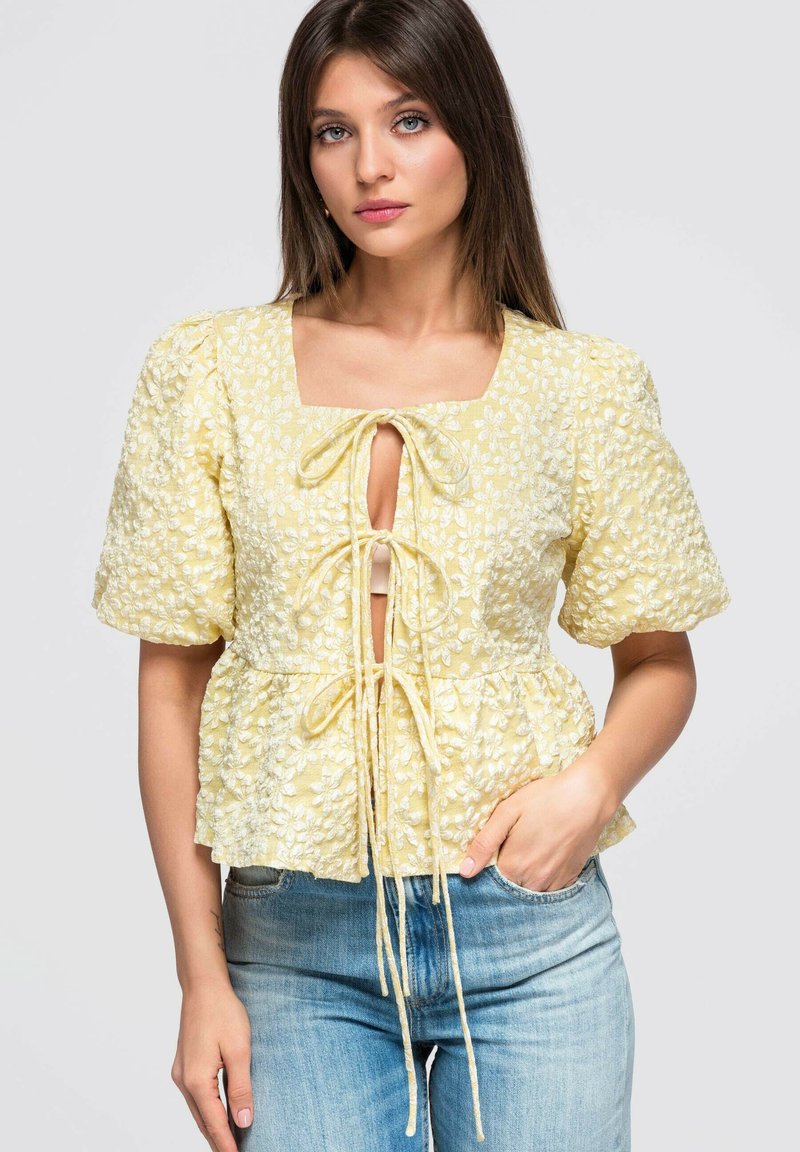 Femme portant un chemisier jaune texturé à motifs floraux, noué avec trois nœuds à l'avant, assorti à un jean bleu clair.
