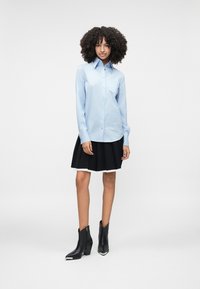 Camisa azul clara de botões com mangas longas, saia preta plissada e botas pretas até o tornozelo com detalhes em prata.