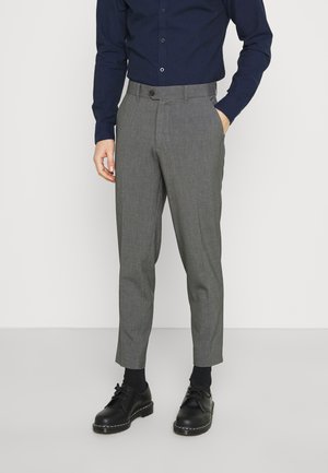 Mann trägt graue maßgeschneiderte Hose, schwarze elegante Schuhe, schwarze Socken und ein dunkelblaues Langarmhemd und steht mit einer Hand in der Tasche.