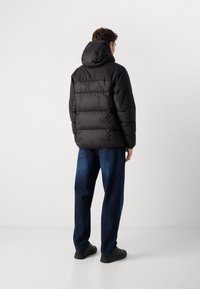 Zwarte gewatteerde jas met een capuchon, voorzien van gesteppete secties en een ontspannen pasvorm, in combinatie met donkerblauwe jeans en zwarte schoenen.