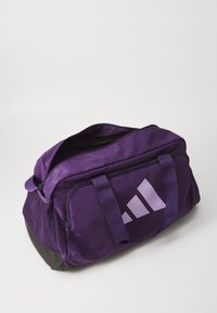 Lilla sports duffel bag med svart bunn, glidelåset hovedrom, to bærehåndtak og stor hvit logo på siden.