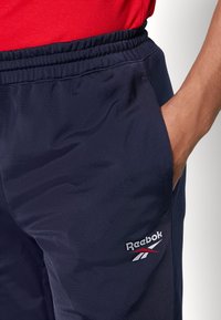 Marineblå sportsbukser med elastisk talje, sidelommer og en hvid Reebok-logo med røde accenter på nederste ben. Blødt, let materiale.