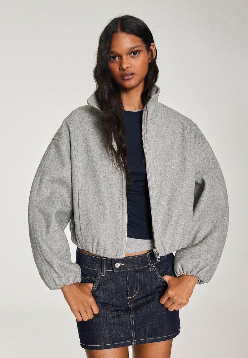 Femme aux longs cheveux foncés portant une veste grise zippée avec des manches bouffantes et une mini-jupe en denim foncé, debout devant un fond uni.