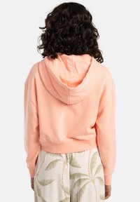 Lichtroze cropped hoodie met een trekkoordcapuchon en geribbelde onderkant; gecombineerd met witte broek met groene palmbladvormen.