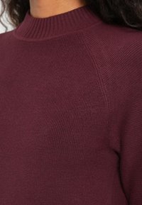 Ingrandimento di una persona che indossa un maglione a coste, con collo alto e maniche lunghe, di colore bordeaux scuro, realizzato in un tessuto lavorato.