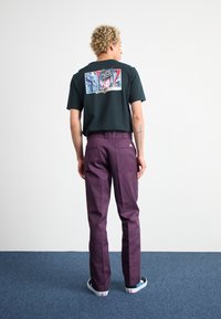 Tricou negru din bumbac cu imprimeu grafic pe spate, combinat cu pantaloni violet și pantofi negri cu accente turcoaz.