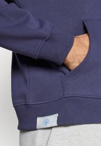 Sudadera navy con puños acanalados y bolsillo, que presenta una etiqueta azul clara con un logo en el dobladillo, hecha de una tela suave.