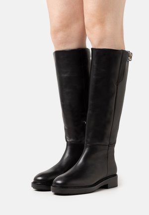 Bottes en cuir noires montant jusqu'aux genoux avec talons blocs bas, portées par une personne aux jambes nues sur fond blanc.