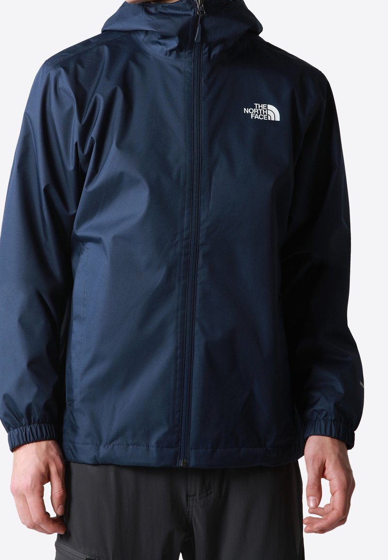 Homme portant une veste de pluie bleu marine zippée avec capuche et le logo "The North Face" sur la poitrine gauche.