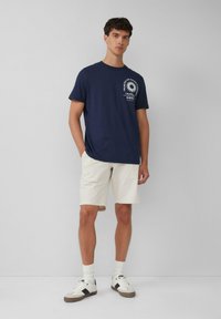 Navyblaue Baumwoll-T-Shirt mit Rundhalsausschnitt und weißem Grafikdruck, kombiniert mit beigen Shorts. Das Model steht vor einem neutralen Hintergrund.