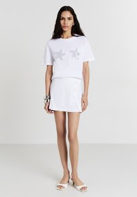 T-shirt blanc avec des embellissements en étoile de mer argentée, associé à une jupe blanche avec des boutons sur le côté et une légère fente. Tissu lisse, design décontracté.
