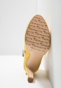 Chaussure à talon jaune inclinée pour montrer la semelle avec motif répétitif du logo "tamaris" et indication de la taille 37 sur fond blanc.