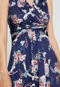 Marineblaues Blumenkleid mit Wickeldesign, das pinke und weiße Blumen, blaue Akzente und einen strukturierten Stoff aufweist. Sichtbares gerafftes Taillendetail.