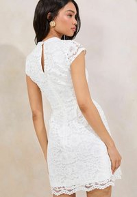 Weißes Spitzenkleid mit Ärmelchen, tailliertem Oberteil und einer Schlüssel-Ausschnitt-Verschluss auf der Rückseite. Der gewellte Saum verleiht Textur und Designinteresse.