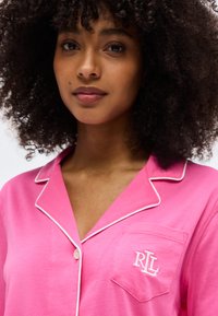 Νεαρή γυναίκα με σγουρά μαλλιά, φορώντας ροζ πουκάμισο με κουμπιά και λευκή διακοσμητική ραφή, με τσέπη κεντημένη με τα γράμματα „R L“.