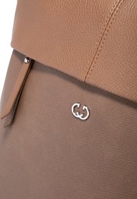 Bolso de cuero marrón texturizado con una solapa suave, detalles en hardware plateado y un logo prominente. Presenta un bolsillo con cremallera y costuras sutiles.