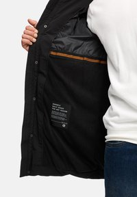 Chaqueta negra exterior con un forro interior suave y textura de polar. Presenta una franja naranja y una etiqueta de cuidado en el interior.