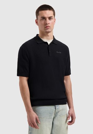 Jonge man met kort haar, gekleed in een zwart getextureerd polohemd met korte mouwen en lichtblauwe versleten spijkerbroek, staand tegen een effen lichte achtergrond.