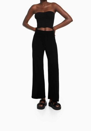 Femme portant un crop top noir côtelé sans bretelles, un pantalon noir large taille haute et des sandales à plateforme noires, debout les mains sur les hanches.