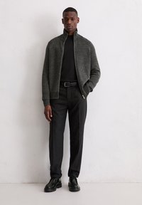 Helaas gebreide zip-up jas in houtskool, zwart lang mouwen shirt, grijze gestructureerde broek en zwarte schoenen. Heeft een staande kraag en ribbelaccenten.