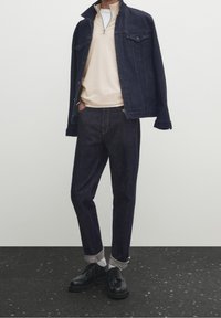 Person wearing dark blue denim jacket draped over shoulders, beige zip-neck sweater, dark rolled-up jeans, white socks, and black shoes.