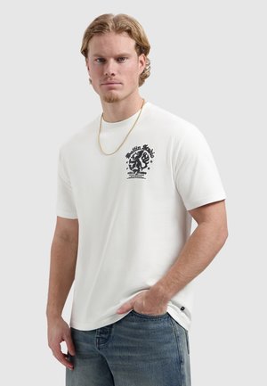 Jonge man met licht haar die een wit T-shirt met opdruk, een goudkleurige ketting en een blauwe spijkerbroek draagt, staand met één hand in zijn zak.