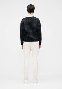 Camisola preta texturizada com padrão geométrico, punhos e barra. Combinada com calças de treino creme e sapatilhas brancas, capturada de costas.