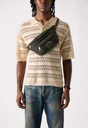 Beige gestricktes Poloshirt mit offenem Muster, getragen über ausgewaschenen blauen Jeans. Grüne Umhängetasche mit Reißverschlüssen und Logo ruht auf der Schulter.