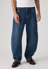 Χαλαρά μπλε τζιν με ψηλή μέση, ραφές σε αντίθεση και κλασικό σχέδιο με πέντε τσέπες, συνδυασμένα με σκούρα μοκασίνια.