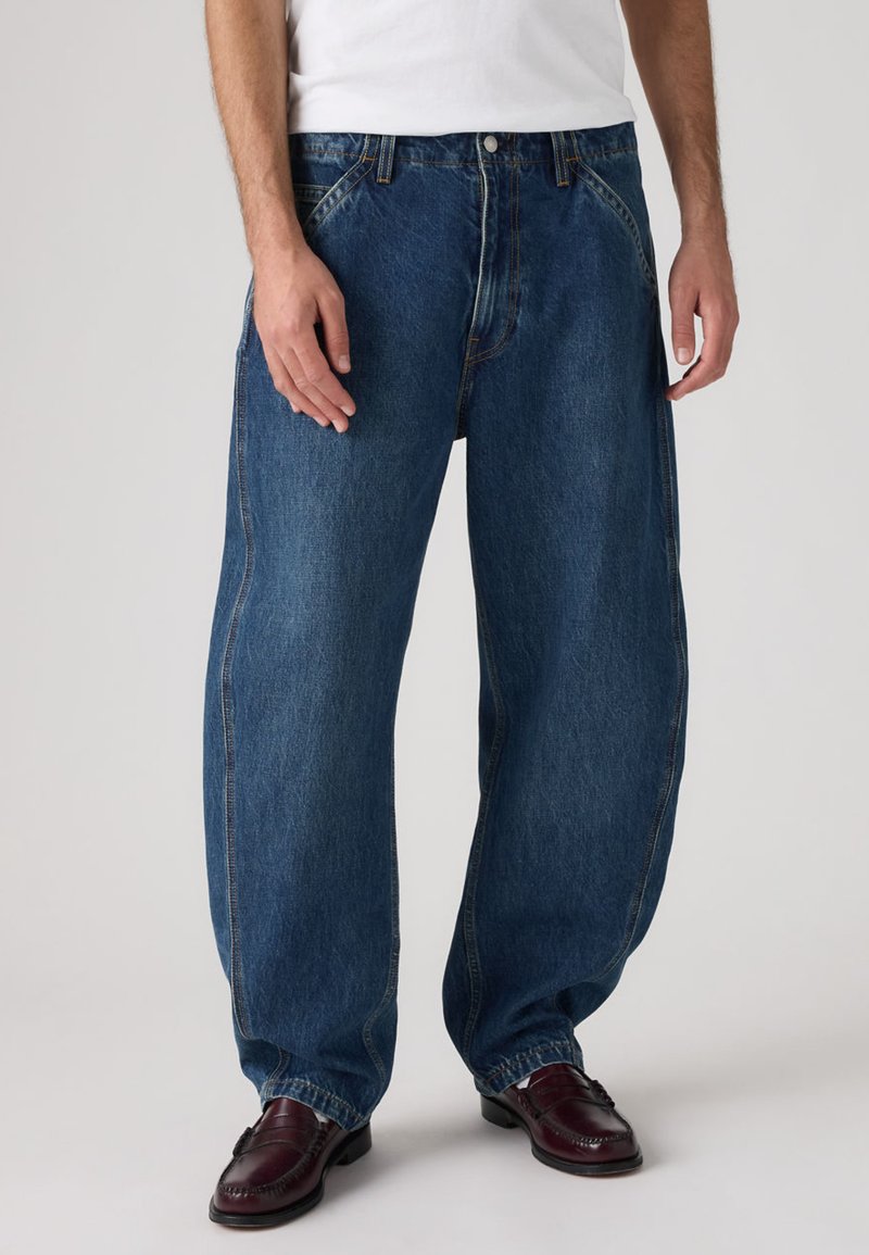 Χαλαρά μπλε τζιν με ψηλή μέση, ραφές σε αντίθεση και κλασικό σχέδιο με πέντε τσέπες, συνδυασμένα με σκούρα μοκασίνια.
