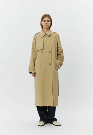 Woman stands wearing a long beige trench coat over dark pants and white shoes against a plain light gray background.