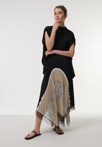 Top negro con mangas oversized combinado con una falda plisada asimétrica que presenta tonos beige, detalles de encaje y acentos negros, llevada con sandalias negras.
