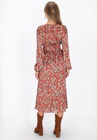 Rochie maxi cu imprimeu roșu și verde, cu mâneci lungi, talie strânsă și siluetă fluidă, asortată cu bocanci din suede maro până la glezne.