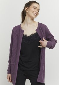 Paarse gebreide cardigan met een ronde halslijn, lange mouwen en zakken, gelaagd over een zwarte top met kantafwerking.