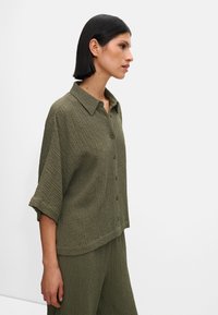 Oliwkowa, krótko rękawowa koszula zapinana na guziki, z teksturowanej tkaniny i z kołnierzykiem. Luźny krój, lekko skrócony dół, z widocznymi guzikami.