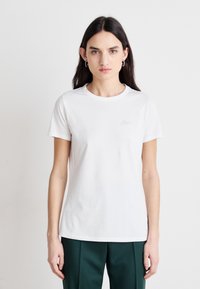 Camiseta blanca de algodón con cuello redondo y mangas cortas, que presenta un pequeño logo gris claro en el lado izquierdo del pecho, combinada con pantalones verdes.