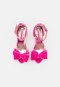 Paire d'escarpins ouvertures à l'avant en velours rose vif avec brides à la cheville et grands nœuds sur le devant, vue de dessus sur fond blanc.