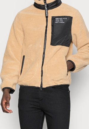 Veste en polaire beige avec poche poitrine zippée noire et fermeture éclair, portée par une personne avec une main dans la poche et un pantalon noir visible.