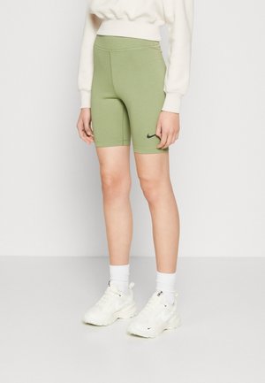 Personne portant un short de vélo Nike vert clair, un sweatshirt blanc, des chaussettes Nike blanches et des baskets Nike blanches, debout sur un sol gris clair.