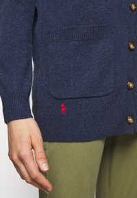 Cárdigan azul marino hecho de un tejido de punto texturizado que presenta botones marrones, un bolsillo en el pecho y un pequeño logo rojo cerca del bolsillo.
