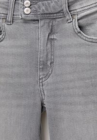 Detailný pohľad na prednú časť šedých riflí so zlátavými gombíkmi, pútkami na opasok, prednými vreckami a viditeľnými švovými detailmi.